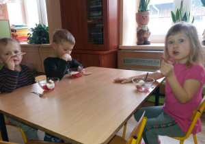 Dwóch chłopców i dziewczynka siedzą przy stoliku i jedzą galaretkę z bitą śmietaną i wafelka w kształcie serca